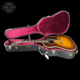 An acoustic guitar with a natural wood finish resting inside an open black hard case with a magenta-pink velvet interior, shown against a solid black background. A Tone Shop watermark is visible in the top-left corner.