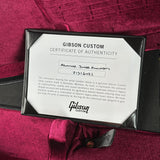 A close-up of a Gibson Custom Certificate of Authenticity card inside a black frame, placed on a deep magenta velvet surface with part of a guitar visible at the bottom.