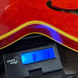 Close-up of a red electric guitar body resting on a black smart weigh scale, showing the scale's digital display reading 8 lbs and 1.3 oz.