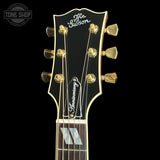 Close-up of a Gibson guitar headstock with gold tuning pegs and strings on a black background. The headstock is black with gold trim and features the Gibson logo at the top and a 'Frontier Century' pickguard in the center.
