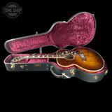 An acoustic guitar with a sunburst finish displayed inside an open black hard case with a magenta velvet interior, set against a solid black background.