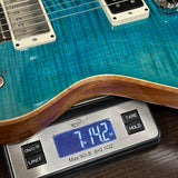 A close-up of a blue electric guitar resting on a digital scale displaying 7.142 lbs, with the guitar's wood body edge and pickups visible.