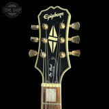 Close-up of an Epiphone Les Paul Custom electric guitar headstock, showing the gold tuners, black and gold headstock design, and part of the fretboard against a black background.