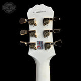 Close-up of a white guitar headstock with six gold-toned tuning pegs and a small red and white sticker near the bottom, photographed against a black background. A Tone Shop watermark is visible in the upper left corner.