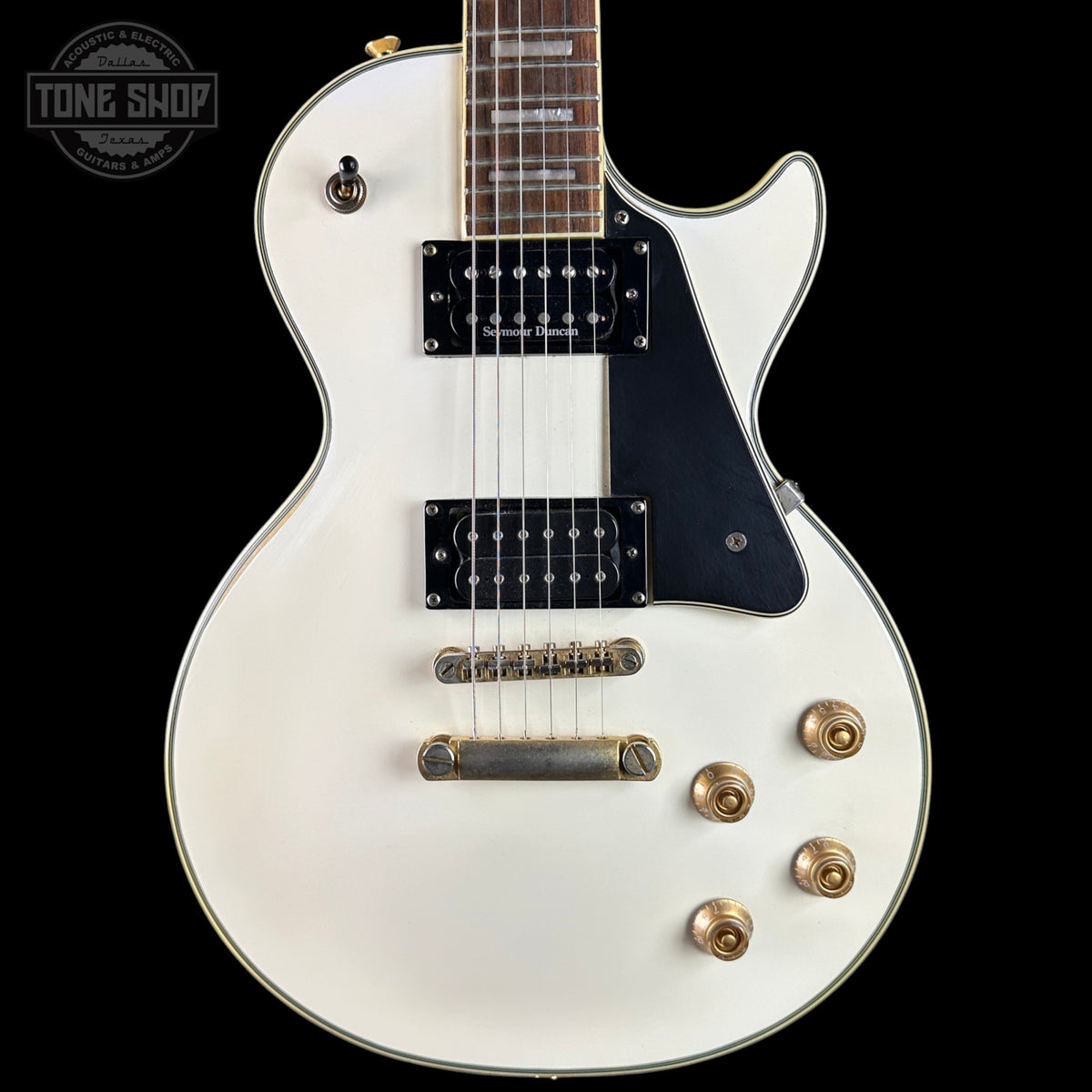 A white electric guitar with a black pickguard and gold hardware, shown from the side against a black background. A Tone Shop watermark is visible in the top-left corner.