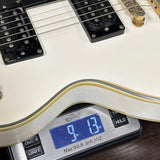 Close-up of a white electric guitar body resting on a digital scale displaying 9.13 oz, with black pickups and part of the neck visible.