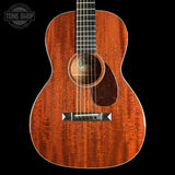 A close-up studio shot of an acoustic guitar body with a warm reddish-brown wood finish, black pickguard, and silver bridge, shown against a solid black background. A Tone Shop watermark is visible in the top-left corner.