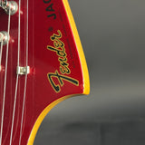 Close-up of a Fender electric guitar headstock in red with a yellow border and black-and-gold Fender logo, showing part of the tuning pegs and strings against a dark gray background.