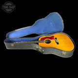 An acoustic guitar with a natural wood finish and red pickguard, shown inside an open blue-lined hard case on a black background. A Tone Shop watermark is visible in the top left corner.