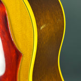 Close-up detail of an acoustic guitar body showing the natural wood back, a gold-toned top, a red headstock edge, and a yellow binding line against a dark gray background.