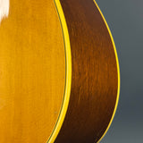 Close-up detail of an acoustic guitar body showing the gold-toned top and dark brown wood sides with a thin light-colored edge trim, set against a dark gray background.