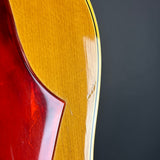 Close-up of a guitar body showing the gold-toned wood top edge and a glossy red-orange sunburst section, photographed against a dark gray background.