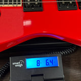 Close-up of a red electric guitar body resting on a Smart Weigh digital scale displaying 8 lbs 6 oz, with the guitar pickups and part of the neck visible above the scale.