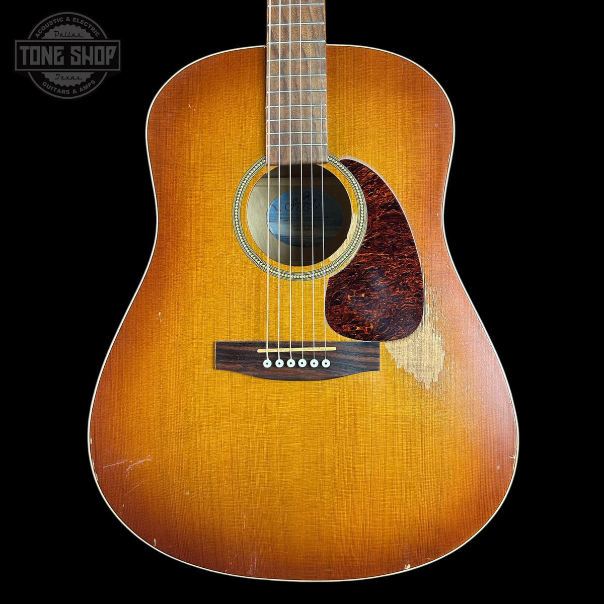 A vintage acoustic guitar with a natural sunburst finish, shown from the front against a solid black background. The guitar has a dark pickguard and a visible wear patch on the lower right side of the body.