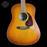 A vintage acoustic guitar with a natural sunburst finish, shown from the front against a solid black background. The guitar has a dark pickguard and a visible wear patch on the lower right side of the body.