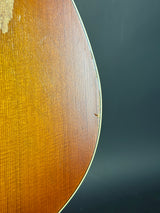 Close-up of the curved wooden body of an acoustic guitar, showing the natural wood grain and a small defect on the top surface, set against a dark gray background.