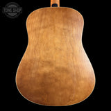 The back of an acoustic guitar made of natural wood with a warm honey-brown finish and visible grain patterns, shown against a solid black background with a Tone Shop watermark in the top left corner.
