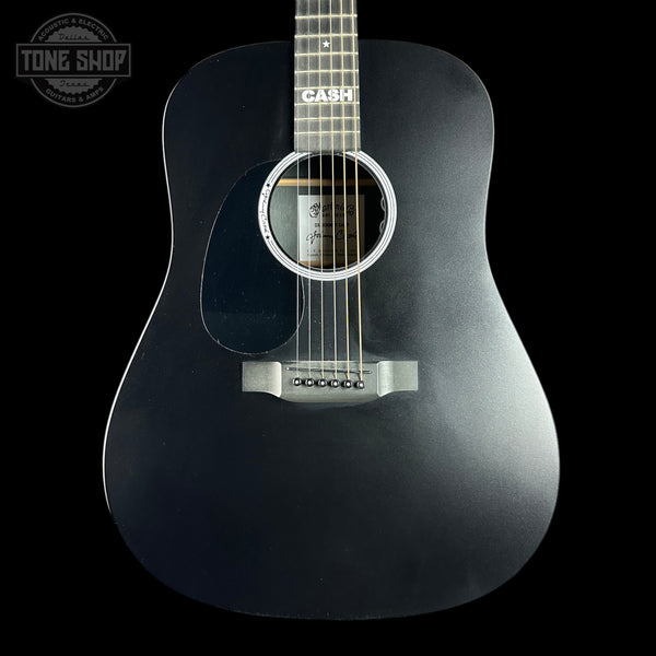 A black acoustic guitar with a matte black finish, featuring a white rosette around the sound hole and a silver bridge, shown against a solid black background. The brand name 'CASH' is printed on the neck.