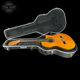 A classical acoustic guitar with a natural wood finish resting inside an open gray hard case, shown against a solid black background. A Tone Shop logo is visible in the top-left corner.