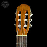 Close-up of a classical guitar headstock made of warm brown wood, with six white tuning keys and strings extending down into the dark fretboard, set against a solid black background. A Tone Shop watermark is visible in the top-left corner.