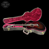 An acoustic guitar with a dark brown sunburst finish displayed inside an open hard case with a bright pink velvet interior, set against a solid black background.