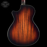 Close-up of the back of an acoustic guitar showing a rich sunburst wood finish that transitions from bright amber-orange in the center to dark brown and black at the edges, set against a solid black background with a Tone Shop watermark in the top-left corner.