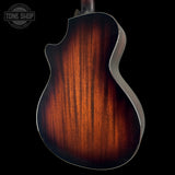 Close-up of the back of an acoustic guitar showing a rich sunburst finish that transitions from bright amber-orange in the center to deep black around the edges, set against a solid black background with a Tone Shop watermark in the top-left corner.