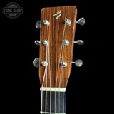 Close-up of an acoustic guitar headstock with a warm brown wood finish, six silver tuning pegs, and six strings, shown against a solid black background with a Tone Shop watermark in the top-left corner.