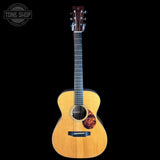 A natural wood acoustic guitar with a dark headstock, shown vertically against a solid black background. A circular 'TONE SHOP' watermark is visible in the top-left corner.