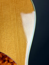 Close-up of a guitar body showing a light natural wood top with a glossy finish, a dark navy background, and part of a darker brown wood inlay in the lower right corner.