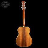 The back of a wooden acoustic guitar, showing a rich grain pattern in warm brown tones, photographed against a solid black background with a Tone Shop watermark in the top-left corner.