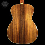 Close-up of the back of an acoustic guitar showing a rich, warm brown wood grain finish, set against a solid black background with a Tone Shop watermark in the upper left.