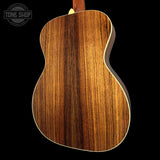 Close-up of the back of an acoustic guitar with a rich, dark brown wood grain finish and a cream-colored edge trim, shown against a solid black background with a Tone Shop watermark in the top left corner.