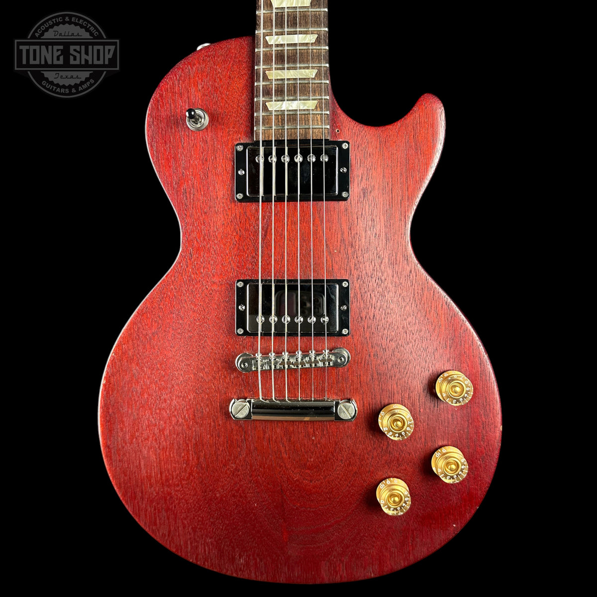 A red electric guitar with a glossy red finish, black pickups, and gold-toned control knobs, shown against a solid black background with a Tone Shop watermark in the top left corner.