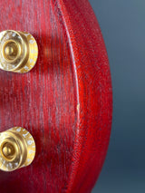 Close-up of a glossy dark red wooden guitar headstock showing the grain of the wood and part of two gold-toned tuning pegs against a dark gray background.