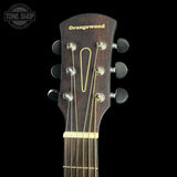 Close-up of an acoustic guitar headstock made of dark brown orangewood, featuring gold lettering, six tuning pegs, and a gold truss rod cover, set against a solid black background.