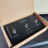 Black guitar effects pedal with 'Boost', 'Reverb', and 'Tremolo' switches in an open cardboard box.