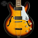 A close-up of an electric guitar body against a black background, featuring a sunburst finish that transitions from bright amber-yellow in the center to dark brown and black at the edges. It has a white pickguard with a stylized 'e' logo, dual humbucker pickups, gold-toned hardware, and a chrome tailpiece.