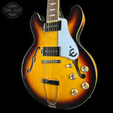 A classic double-cutaway electric guitar with a sunburst finish, featuring a white pickguard with a black logo, gold-toned hardware, and a dark fretboard, shown against a solid black background.