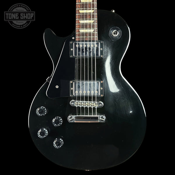A black electric guitar with chrome hardware, shown from the side against a black background. A circular 'TONE SHOP' watermark is visible in the top-left corner.