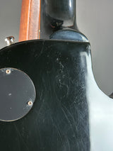 Close-up of the glossy black back of an electric guitar body, showing visible scratches and a circular mounting plate with screws, with a brown wood neck visible on the left.