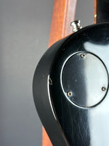Close-up detail of a glossy black guitar body showing a circular plate secured by four screws, with part of a wooden neck and a metal control visible.