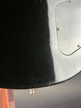 Close-up of a glossy black guitar body with a silver pickguard and a small metal screw on the right side, photographed indoors against a gray background.