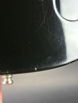 Close-up of a black glossy guitar body showing surface scratches and a small silver hardware piece against a gray background.