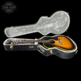 A vintage acoustic guitar with a sunburst finish resting inside an open black hard case with gray padding, shown against a solid black background. A Tone Shop logo is visible in the top left corner.