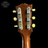 Close-up of the headstock of a vintage acoustic guitar, showing the brown wood grain, metal tuning machines, and cream-colored tuning pegs against a solid black background.