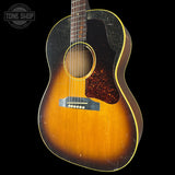A vintage acoustic guitar with a sunburst finish, featuring a black pickguard, a dark fretboard, and a gold-toned binding, shown against a solid black background.