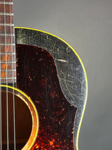 Close-up of an acoustic guitar body showing a heavily scratched black pickguard over a dark tortoiseshell-style faceplate, with a yellow binding and part of the fretboard visible against a gray background.