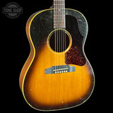 A vintage acoustic guitar with a classic sunburst finish, featuring a dark black pickguard and a dark fretboard, shown against a solid black background with a Tone Shop watermark in the top left corner.
