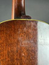 Close-up of the back of a wooden acoustic guitar showing the rich brown wood grain, a yellow binding edge, and a worn, scuffed varnish on the right side.